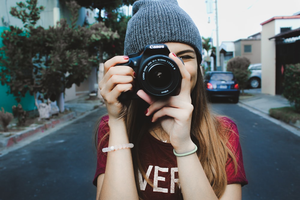 woman wearing red t-shirt and gray knitted cap standing on concrete road using Canon bridge camera during daytime