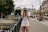 A person wearing a cap and casual clothing stands on a city street, holding a camera and taking a photo. The background features historic European architecture, with people sitting on benches and walking nearby. Trees and a bridge railing are visible, suggesting an urban setting.