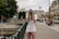 A person wearing a cap and casual clothing stands on a city street, holding a camera and taking a photo. The background features historic European architecture, with people sitting on benches and walking nearby. Trees and a bridge railing are visible, suggesting an urban setting.