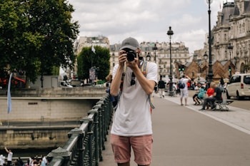 A person wearing a cap and casual clothing stands on a city street, holding a camera and taking a photo. The background features historic European architecture, with people sitting on benches and walking nearby. Trees and a bridge railing are visible, suggesting an urban setting.