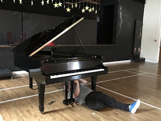 A grand piano is situated in a room with a wooden floor and a black backdrop adorned with hanging stars. A person is lying on the floor underneath the piano, possibly engaged in some kind of repair or maintenance work. The scene has a casual and slightly humorous feel due to the unusual positioning of the person.