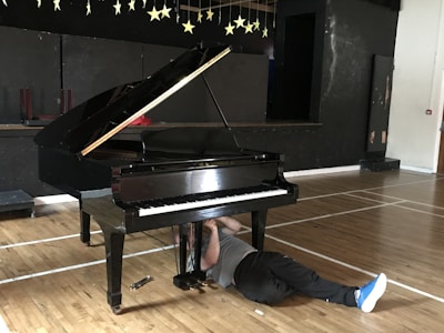 A grand piano is situated in a room with a wooden floor and a black backdrop adorned with hanging stars. A person is lying on the floor underneath the piano, possibly engaged in some kind of repair or maintenance work. The scene has a casual and slightly humorous feel due to the unusual positioning of the person.