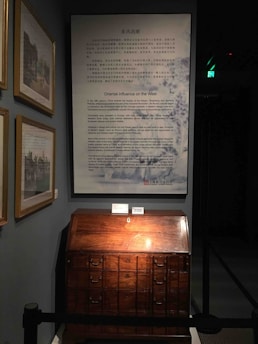 A museum exhibit features a vintage wooden cabinet with several drawers, illuminated by overhead lighting. Above the cabinet, a large informational poster titled 'Oriental Influence on the West' is displayed, providing historical context. The setting includes framed pictures on the wall to the left and a dark background contributing to the focused lighting on the exhibit.