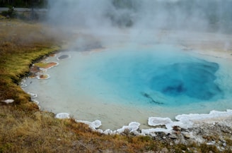 A geothermal hot spring with a vivid blue center surrounded by white mineral deposits and multicolored edges. Mist rises from the water, creating a mysterious and tranquil atmosphere against the backdrop of grassy terrain.