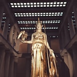 A large golden statue of a goddess stands in an elaborate temple setting. The goddess holds a small figure in one hand and a shield in the other. The scene is framed by tall columns and a patterned ceiling, evoking a sense of grandeur and classical architecture.