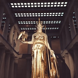 A large golden statue of a goddess stands in an elaborate temple setting. The goddess holds a small figure in one hand and a shield in the other. The scene is framed by tall columns and a patterned ceiling, evoking a sense of grandeur and classical architecture.