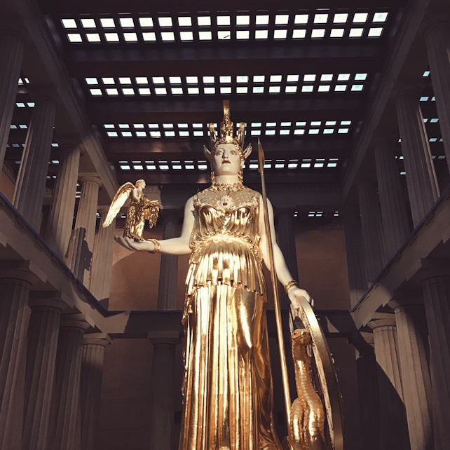 A large golden statue of a goddess stands in an elaborate temple setting. The goddess holds a small figure in one hand and a shield in the other. The scene is framed by tall columns and a patterned ceiling, evoking a sense of grandeur and classical architecture.
