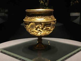 An ornate golden goblet with intricate engravings featuring winged creatures, possibly griffins, and decorative patterns. It is placed on a glass display under focused lighting, highlighting its details and craftsmanship.