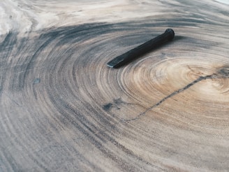 A large, textured wooden surface with visible rings, similar to tree growth rings, with a single rusty nail laid flat across the pattern. The wood has a natural gradient of light to dark brown tones, highlighting the intricate grain and natural beauty of the material.