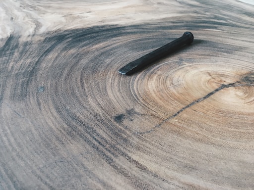 A large, textured wooden surface with visible rings, similar to tree growth rings, with a single rusty nail laid flat across the pattern. The wood has a natural gradient of light to dark brown tones, highlighting the intricate grain and natural beauty of the material.