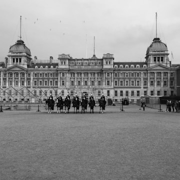 A large historical building with ornate architecture is prominently featured in the background. In the foreground, a formation of mounted guards is present, dressed in traditional attire. The scene appears to be a formal event or ceremony, with a few people observing from the sidelines. The sky is overcast, adding a somber tone to the setting.