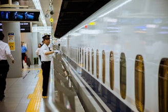A train conductor in a white uniform and cap stands beside a sleek white bullet train, gesturing with a white-gloved hand. The setting is a modern train station with passengers in the background, some carrying luggage. Overhead signage is visible, including directions and escalator signs.