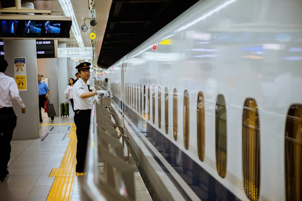 A train conductor in a white uniform and cap stands beside a sleek white bullet train, gesturing with a white-gloved hand. The setting is a modern train station with passengers in the background, some carrying luggage. Overhead signage is visible, including directions and escalator signs.