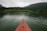 A red canoe or kayak is floating on a calm, greenish body of water surrounded by lush greenery. Dense vegetation lines the shores on either side and distant hills are visible under an overcast sky, creating a peaceful and secluded atmosphere.