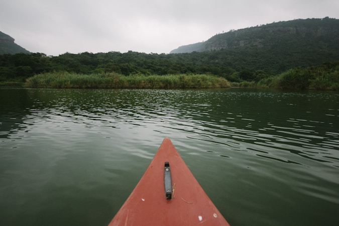 A red canoe or kayak is floating on a calm, greenish body of water surrounded by lush greenery. Dense vegetation lines the shores on either side and distant hills are visible under an overcast sky, creating a peaceful and secluded atmosphere.
