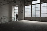 A woman in a floral dress stands near a large window in an empty, dimly lit room with tiled floors. The space features industrial-style architecture and large glass-paneled doors that allow light to filter in, casting a soft glow. A 'descida' sign indicates an exit, adding to the ambiance of solitude.