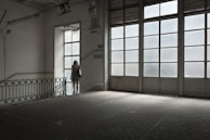 A woman in a floral dress stands near a large window in an empty, dimly lit room with tiled floors. The space features industrial-style architecture and large glass-paneled doors that allow light to filter in, casting a soft glow. A 'descida' sign indicates an exit, adding to the ambiance of solitude.