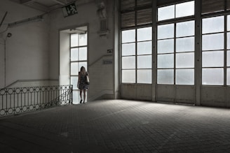 A woman in a floral dress stands near a large window in an empty, dimly lit room with tiled floors. The space features industrial-style architecture and large glass-paneled doors that allow light to filter in, casting a soft glow. A 'descida' sign indicates an exit, adding to the ambiance of solitude.