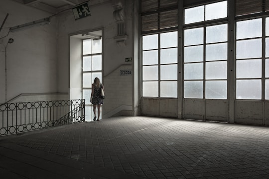 A woman in a floral dress stands near a large window in an empty, dimly lit room with tiled floors. The space features industrial-style architecture and large glass-paneled doors that allow light to filter in, casting a soft glow. A 'descida' sign indicates an exit, adding to the ambiance of solitude.