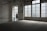 A woman in a floral dress stands near a large window in an empty, dimly lit room with tiled floors. The space features industrial-style architecture and large glass-paneled doors that allow light to filter in, casting a soft glow. A 'descida' sign indicates an exit, adding to the ambiance of solitude.
