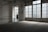 A woman in a floral dress stands near a large window in an empty, dimly lit room with tiled floors. The space features industrial-style architecture and large glass-paneled doors that allow light to filter in, casting a soft glow. A 'descida' sign indicates an exit, adding to the ambiance of solitude.