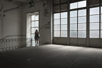 A woman in a floral dress stands near a large window in an empty, dimly lit room with tiled floors. The space features industrial-style architecture and large glass-paneled doors that allow light to filter in, casting a soft glow. A 'descida' sign indicates an exit, adding to the ambiance of solitude.