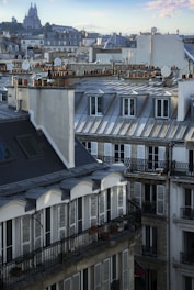 The image captures a scenic view of Parisian rooftops with chimney stacks, satellite dishes, and intricate balconies. The iconic Sacré-Cœur Basilica is visible in the background, perched on a hill. The overall architecture showcases the classic Parisian style with slate roofs and cream-colored facades.