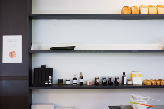 A minimalistic white wall with dark shelves adorned with several loaves of bread on the top shelf, various condiments including jars and bottles neatly arranged on the middle shelf, and baking tools like sifters on the bottom counter. A small sign featuring a bread image is visible to the side.