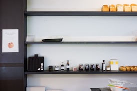A minimalistic white wall with dark shelves adorned with several loaves of bread on the top shelf, various condiments including jars and bottles neatly arranged on the middle shelf, and baking tools like sifters on the bottom counter. A small sign featuring a bread image is visible to the side.