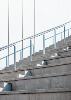 Concrete stairs ascend alongside a glass railing, with sunlight casting geometric shadows on the steps. The sleek, modern design emphasizes clean lines and symmetry.