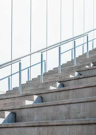 Concrete stairs ascend alongside a glass railing, with sunlight casting geometric shadows on the steps. The sleek, modern design emphasizes clean lines and symmetry.