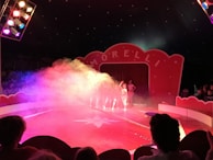 A vibrant circus setting with a stage and large red curtains. The word 'Morelli' is displayed prominently on an arch above the curtains. Colorful stage lights create a mist with hues of pink, purple, and yellow. People are sitting in the audience with silhouettes visible against the illuminated backdrop. Performers are standing in front of the curtains, partially obscured by the colorful haze.