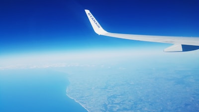 An airplane wing with the logo 'Ryanair' is prominently visible against a backdrop of a clear blue sky. Below, a vast expanse of land and sea is seen, with the coastline distinctly curving from the bottom left corner of the image. There are a few scattered clouds near the horizon.