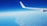 An airplane wing with the logo 'Ryanair' is prominently visible against a backdrop of a clear blue sky. Below, a vast expanse of land and sea is seen, with the coastline distinctly curving from the bottom left corner of the image. There are a few scattered clouds near the horizon.