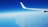 An airplane wing with the logo 'Ryanair' is prominently visible against a backdrop of a clear blue sky. Below, a vast expanse of land and sea is seen, with the coastline distinctly curving from the bottom left corner of the image. There are a few scattered clouds near the horizon.