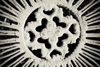 An intricate, circular wrought iron pattern featuring a central ornate design. The image is monochrome, giving it a vintage and textured appearance, with the metalwork casting shadows.