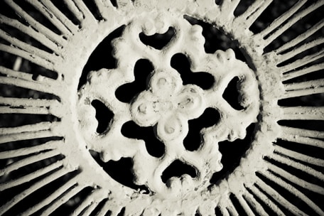 An intricate, circular wrought iron pattern featuring a central ornate design. The image is monochrome, giving it a vintage and textured appearance, with the metalwork casting shadows.