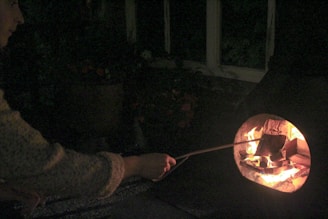 A person is using a stick to tend to a fire in a dark setting. The fire illuminates the interior of a stone or metal fireplace. The ambient environment is dimly lit with minimal visibility of surrounding objects.