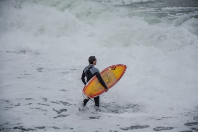 A person wearing a wetsuit is standing on the shore, holding a surfboard with a vibrant red and yellow design. The ocean waves are crashing towards the beach, creating foamy white water around the individual.