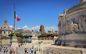 An architectural scene featuring a large circular monument with detailed carvings on the right. In the foreground, a group of people walks across a spacious plaza. A flagpole displays the Italian flag prominently. In the background, historic buildings and a dome structure with classical architecture are visible under a clear blue sky.