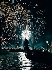A dazzling display of fireworks lights up the night sky over a body of water, with a gondola's silhouette in the foreground. The rippling reflections on the water and a line of illuminated buildings create a festive atmosphere.