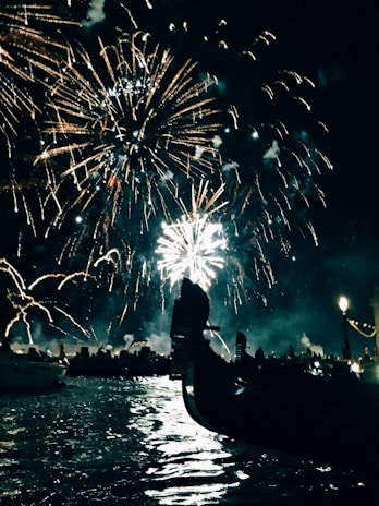 A dazzling display of fireworks lights up the night sky over a body of water, with a gondola's silhouette in the foreground. The rippling reflections on the water and a line of illuminated buildings create a festive atmosphere.