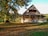 A rustic wooden house with a steep roof is surrounded by a spacious grassy area covered with fallen leaves. Large trees with dense foliage provide shade and a natural backdrop. The warm sunlight casts shadows, creating a peaceful and serene atmosphere.
