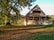 A rustic wooden house with a steep roof is surrounded by a spacious grassy area covered with fallen leaves. Large trees with dense foliage provide shade and a natural backdrop. The warm sunlight casts shadows, creating a peaceful and serene atmosphere.