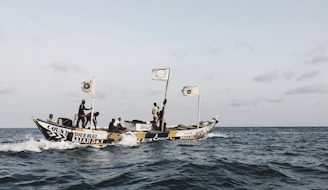 A fishing boat with several people on board moves through the ocean water under a clear sky. The boat is adorned with several flags and colorful text, including phrases like 'Count Your Blessings' and a biblical reference. The people on the boat seem engaged in various activities, possibly related to fishing or traveling.