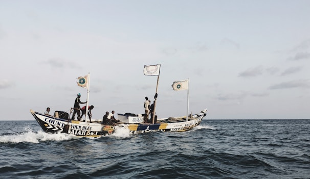A fishing boat with several people on board moves through the ocean water under a clear sky. The boat is adorned with several flags and colorful text, including phrases like 'Count Your Blessings' and a biblical reference. The people on the boat seem engaged in various activities, possibly related to fishing or traveling.