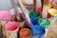 Several open paint cans filled with various bright colors such as pink, blue, green, and red are placed on a surface. Wooden sticks are seen submerged in the paint, likely used for stirring or mixing. Some paint has dripped and splattered around the edges of the cans.