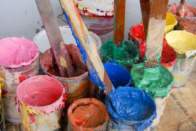 Several open paint cans filled with various bright colors such as pink, blue, green, and red are placed on a surface. Wooden sticks are seen submerged in the paint, likely used for stirring or mixing. Some paint has dripped and splattered around the edges of the cans.