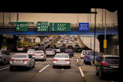 A busy roadway filled with numerous cars, likely during peak traffic hours, beneath a series of blue and green overhead directional signs with Korean and English text. The vehicles are heading towards a bridge while the sky appears overcast.