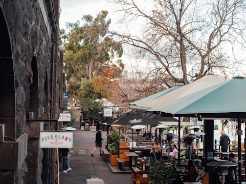 A riverside outdoor dining area with tables protected by large umbrellas. The scene includes groups of people enjoying themselves at the tables, surrounded by trees that are mostly bare except for a few with green leaves. Signs for Riverland and other establishments can be seen along the stone wall of a building.